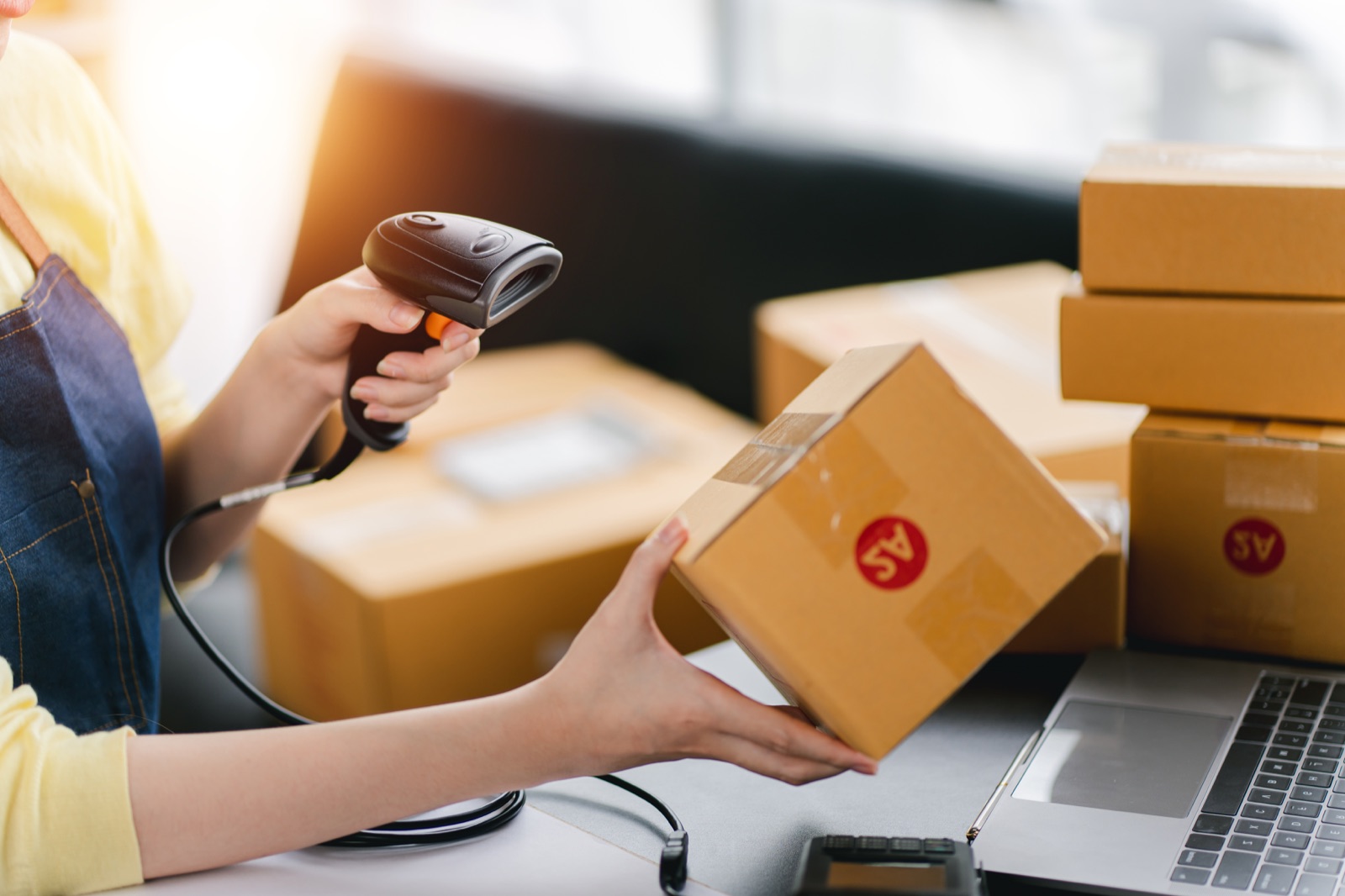 Hände scannen ein Versandpaket mit einem Handscanner, daneben gestapelte Kartons und ein Laptop