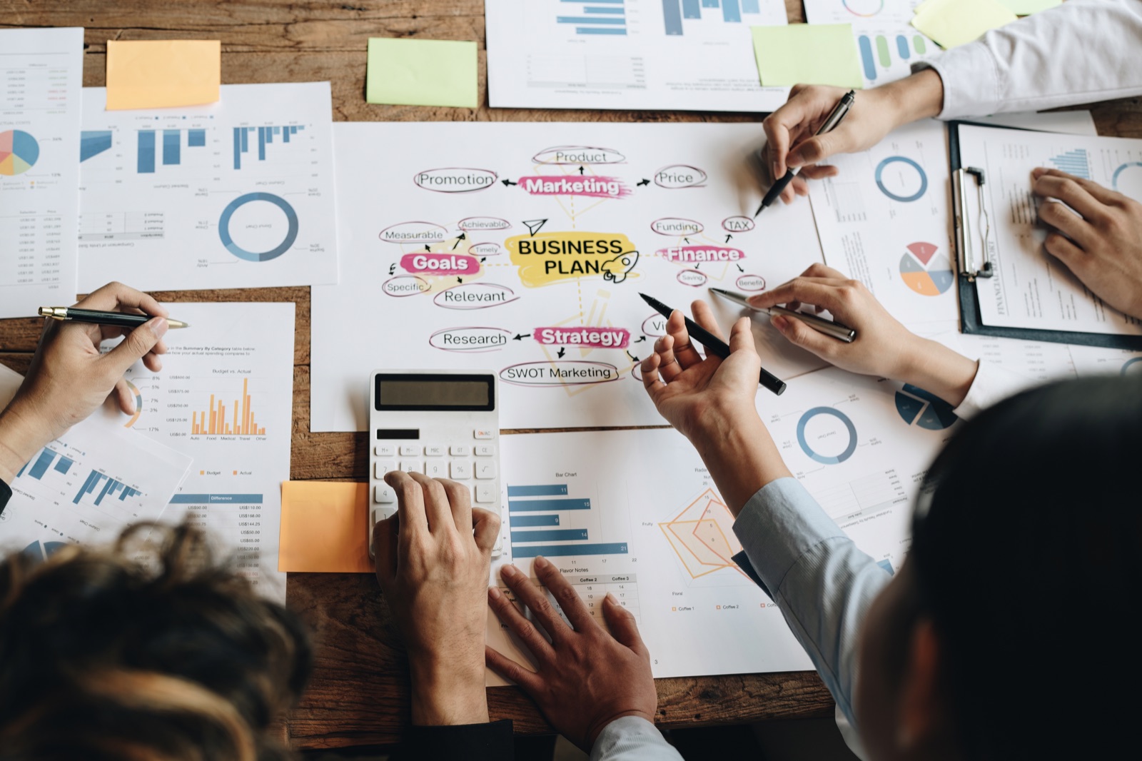 Overhead view of several people at a wooden table with a business plan, charts and notes