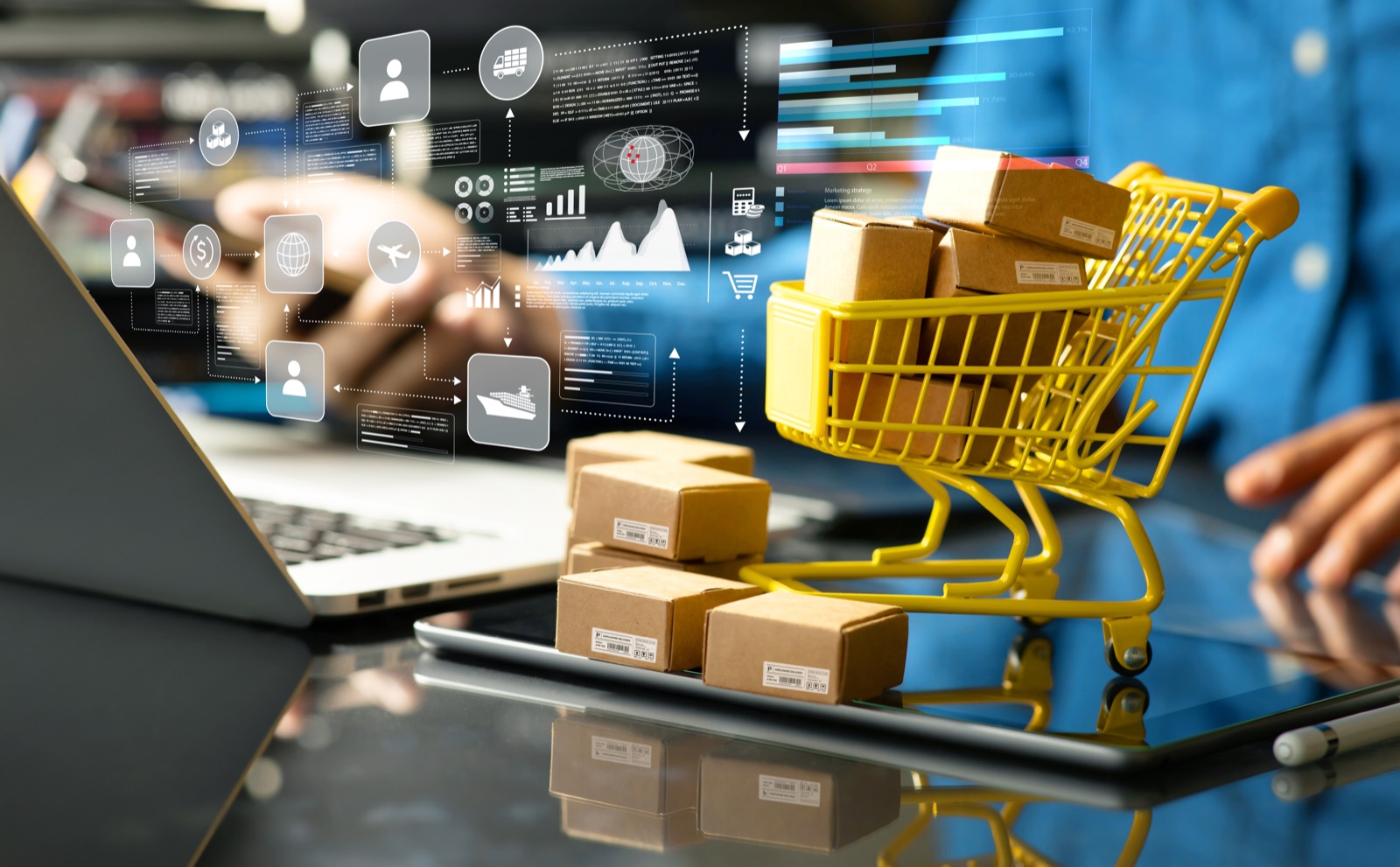 Laptop and tablet with overlaid graphics on orders, shipping and sales, next to a small shopping trolley with parcels